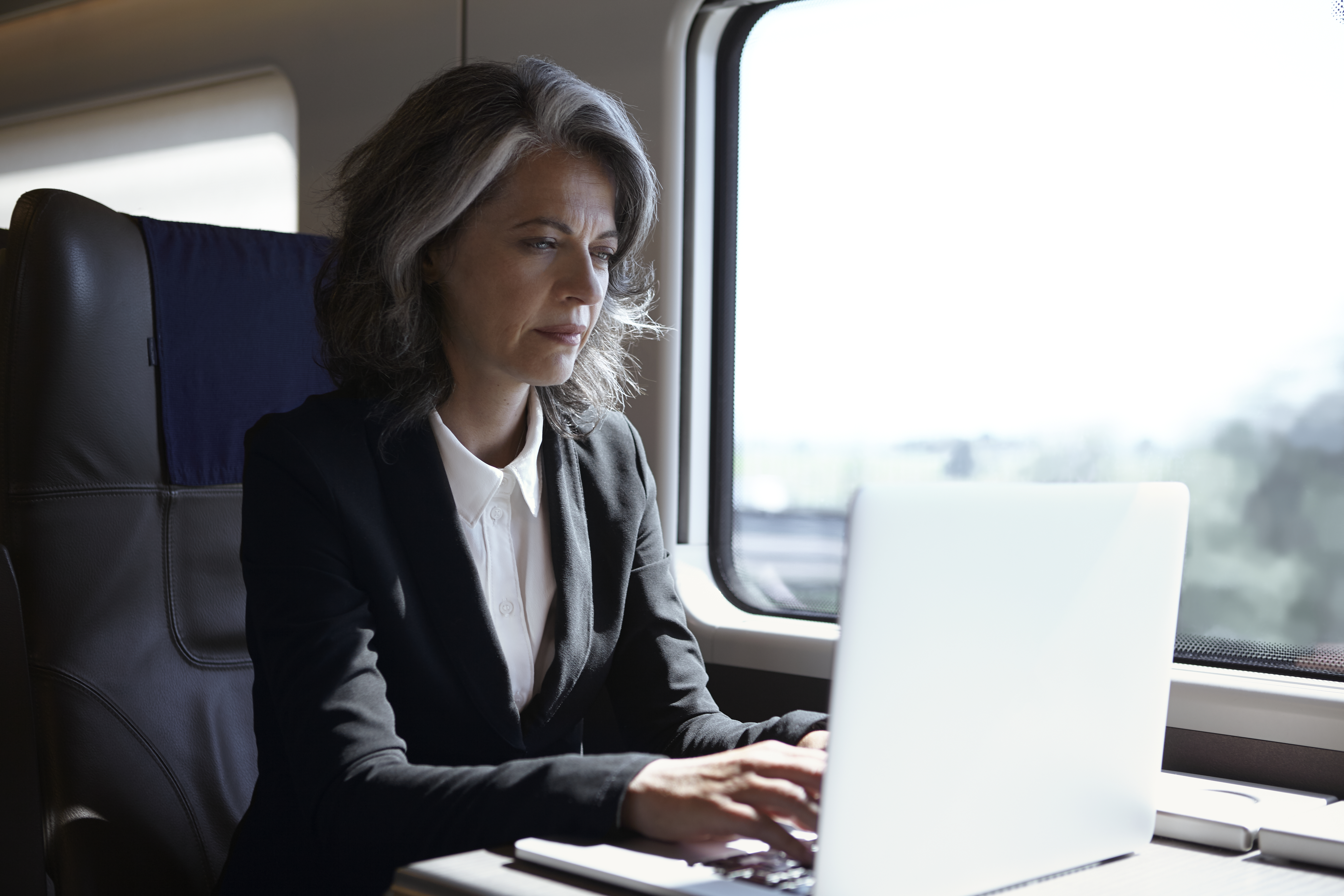 A woman with gray hair, wearing a black blazer and white shirt, types on a laptop while seated on a train near a window. Sunlight illuminates her workspace, and the scenery outside the window is blurred.