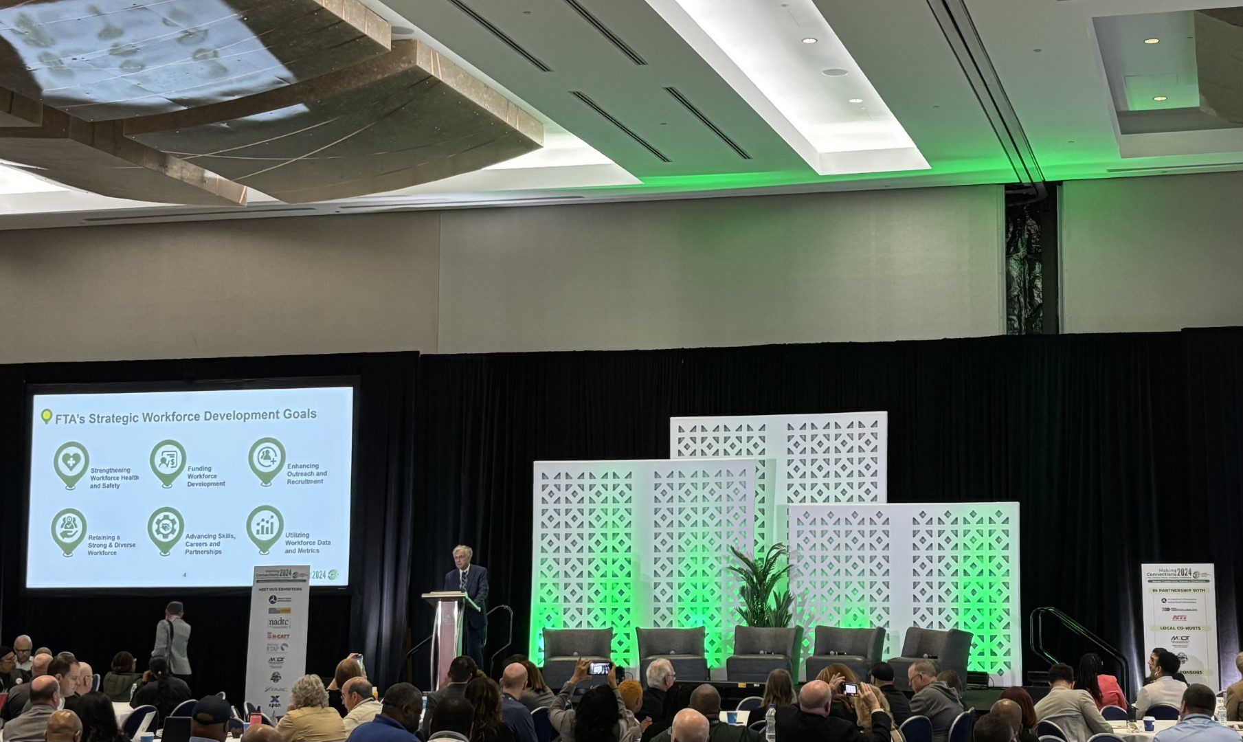 A speaker stands at a podium in a conference hall, delivering a presentation on the FTA's Strategic Workforce Development Goals