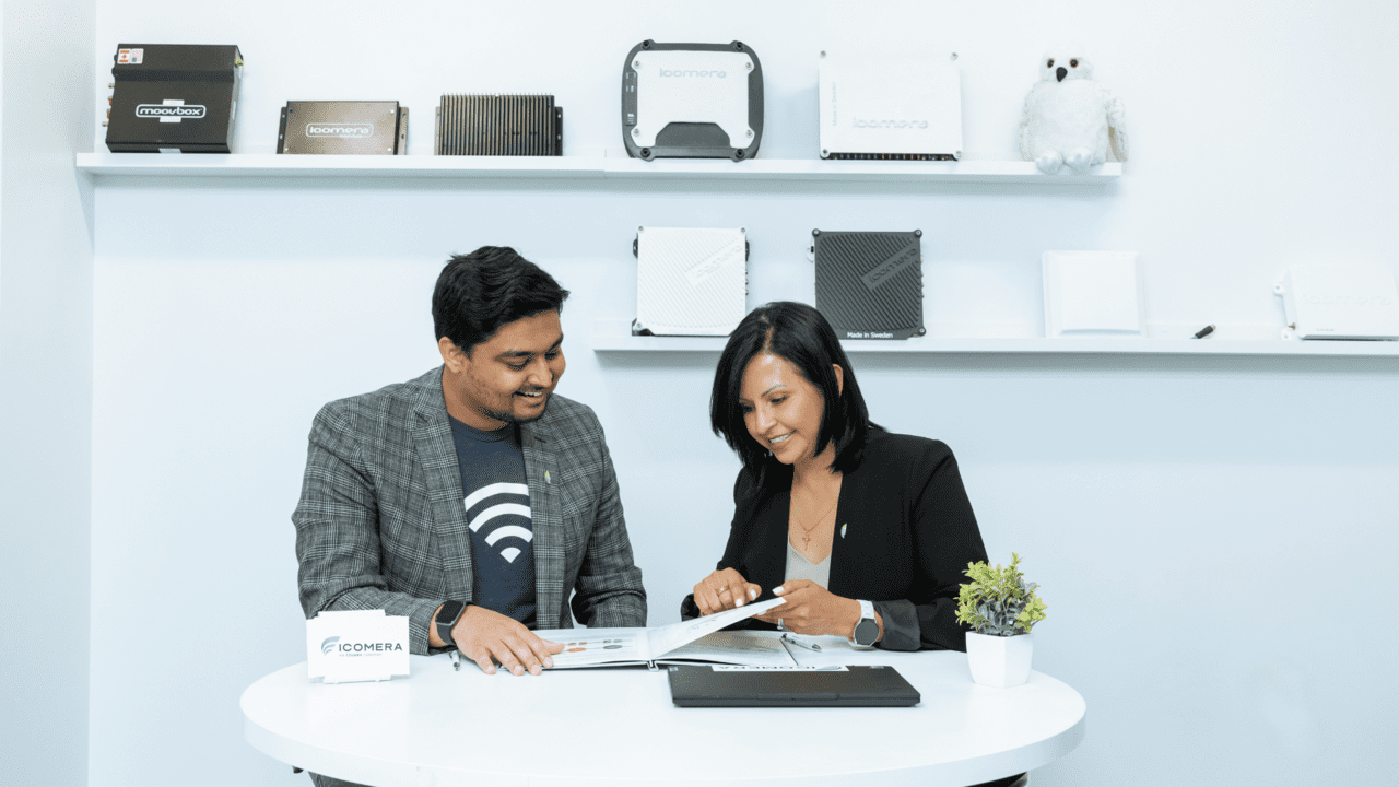 Icomera Senior Project Manager Jackie Morales speaks with a colleague. In the background, Icomera routers are displayed proudly on shelves.